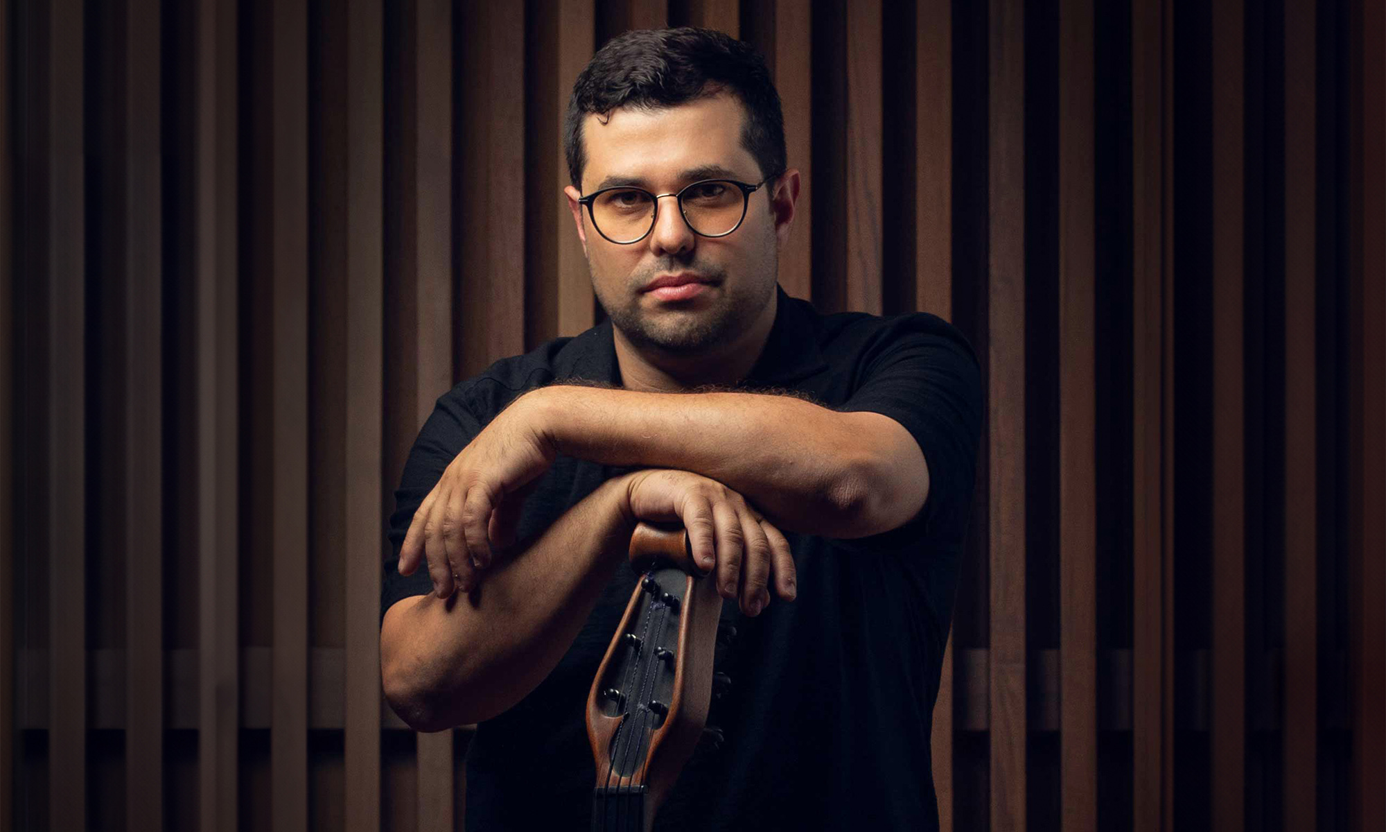 Composer Justin Gray leaning on a bass guitar in front of a brown wood background.